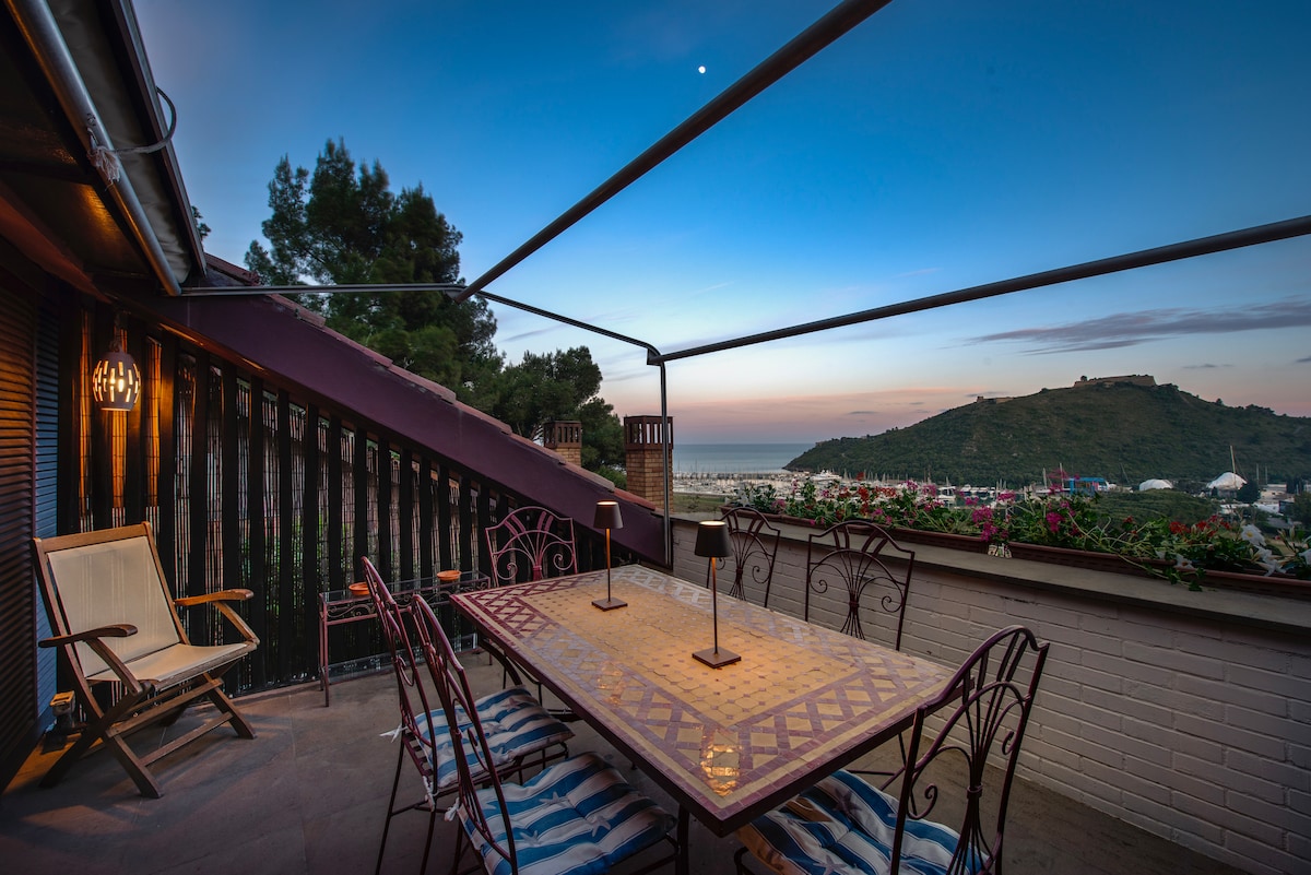 An outdoor terrace is showcased, featuring a large table surrounded by chairs, ideal for dining. The view extends over the sea and hills, with vibrant greenery and flowers visible. A gentle twilight sky provides a serene backdrop.