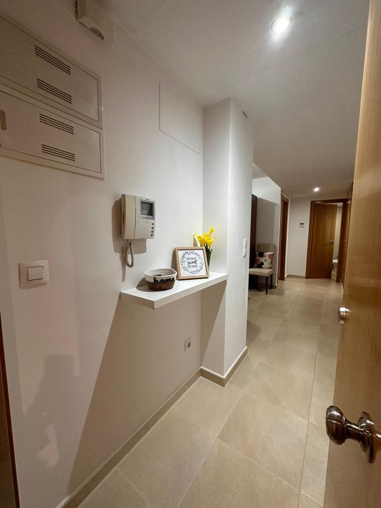 A hallway is shown with a light-colored wall and tiled flooring. A small shelf displays decorative items, including a framed picture and a potted plant. Light fixtures illuminate the space, with doors visible on either side leading to additional rooms.