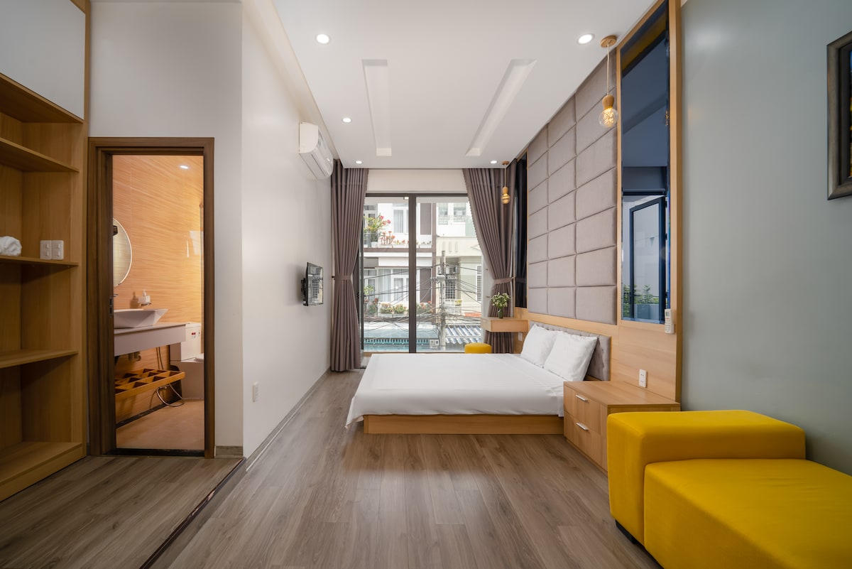A bright bedroom features a spacious bed with white linens against a backdrop of light wood paneling. A comfortable yellow chair sits beside the bed, while large windows allow natural light to fill the room. A modern bathroom is visible through an open door.