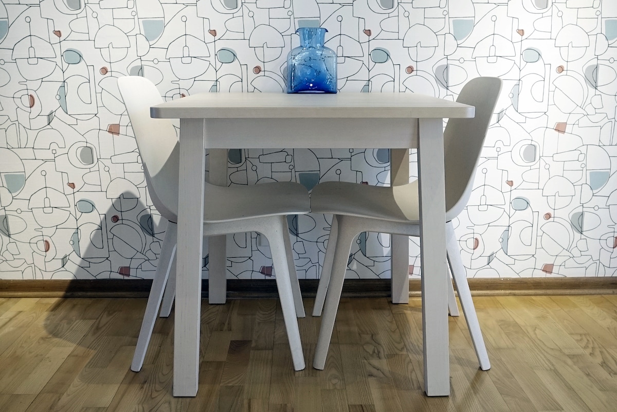 A minimalist dining space is presented with a light-colored table and two white chairs. A striking blue vase sits at the center, adding a pop of color against a patterned wall featuring abstract designs in muted tones.