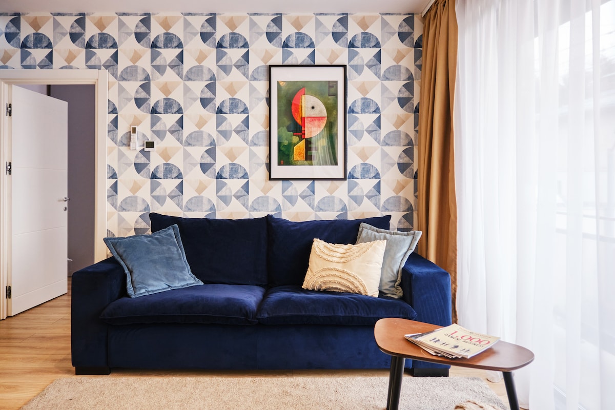 A cozy living area features a dark blue sofa adorned with decorative pillows, complemented by a round wooden coffee table. A patterned wallpaper adds visual interest, while a piece of art hangs above the sofa. Natural light filters through sheer curtains, creating a bright atmosphere.