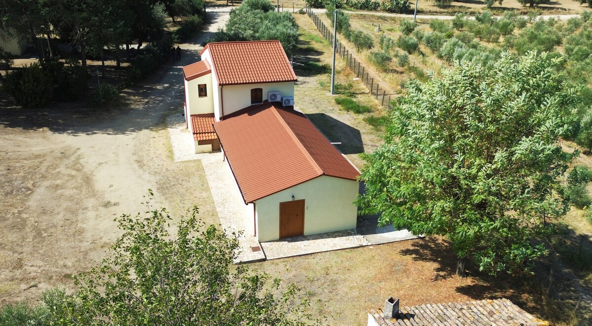 An aerial view captures a two-story building with a terracotta roof and cream-colored walls, surrounded by greenery. The structure is situated within a spacious outdoor area featuring well-maintained paths and trees, providing a serene setting amidst nature.