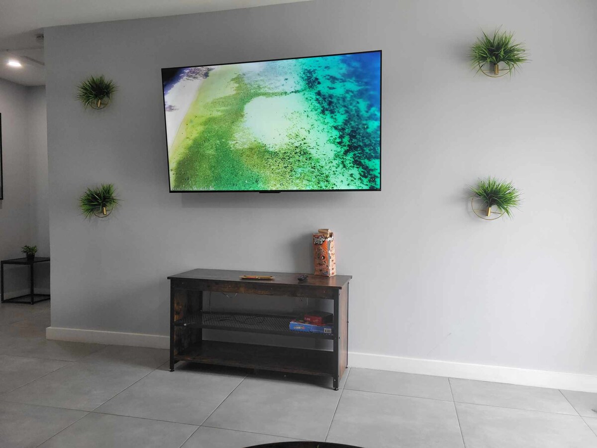 A wall-mounted television displays scenic coastal imagery above a low, wooden console table. Decorative greenery hangs from the wall, complementing the neutral gray backdrop. The space features tiled flooring that enhances the cohesive design.