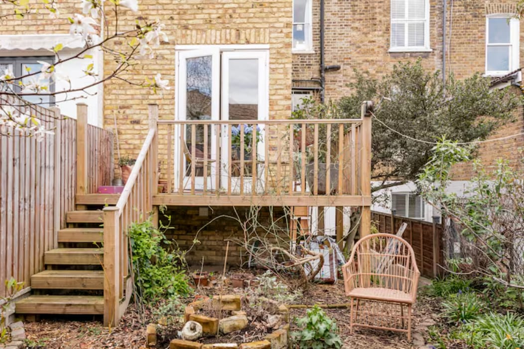 A wooden deck extends from the back of the house, featuring double French doors. Surrounding greenery includes shrubs and small trees, while a decorative chair sits nearby. A set of wooden steps leads down from the deck into the garden.