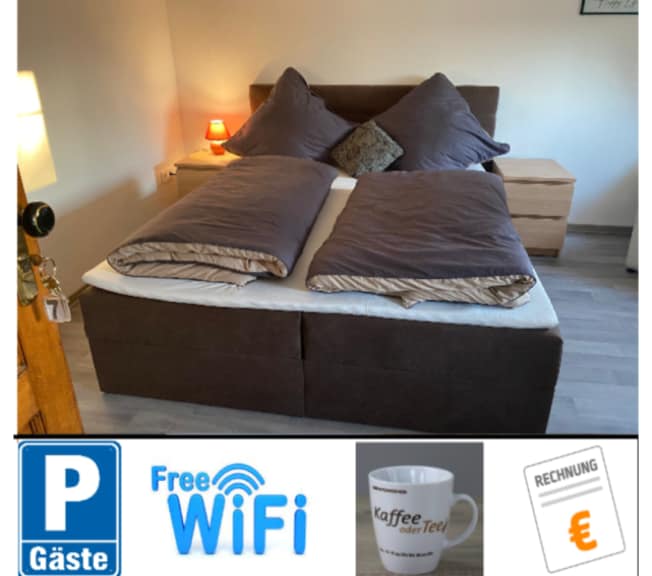 A cozy bedroom features a spacious double bed adorned with two large pillows and a dark-colored duvet. Beside the bed, two wooden nightstands are present. A bedside lamp provides soft lighting, complementing the neutral tones of the room.