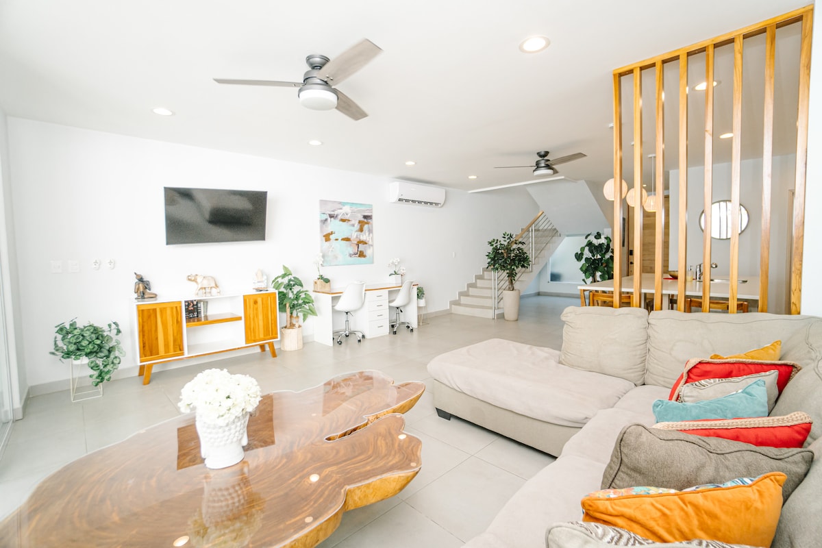 A spacious living area features a modern sofa adorned with colorful cushions, complemented by a unique wooden coffee table. A sleek television is mounted on the wall, while two desks with ergonomic chairs provide workspaces. Plants add a touch of greenery, enhancing the bright atmosphere.