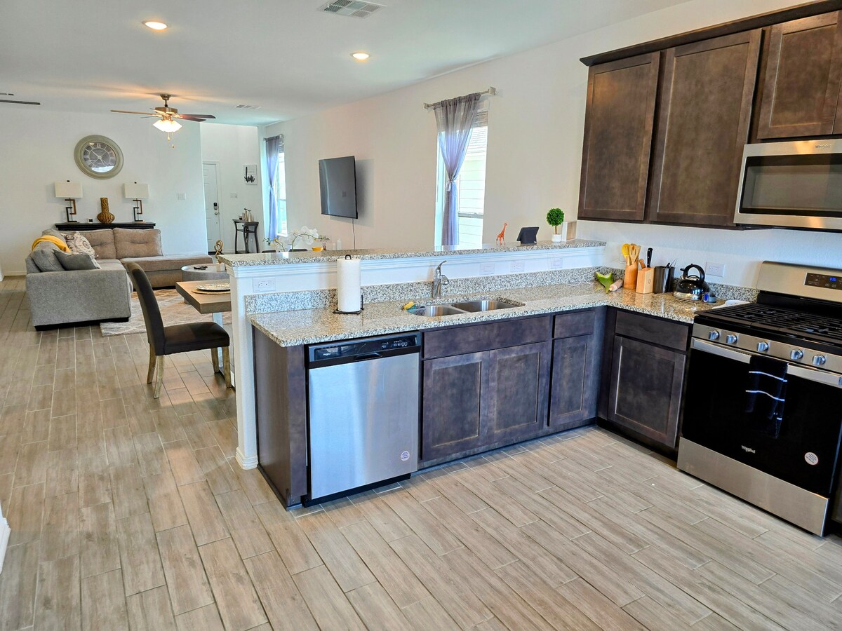 An open kitchen area features modern appliances, including a stainless steel dishwasher and gas range. Countertops provide ample space for meal preparation, while a casual dining area is visible in the background, showcasing a comfortable seating arrangement and natural light from nearby windows.