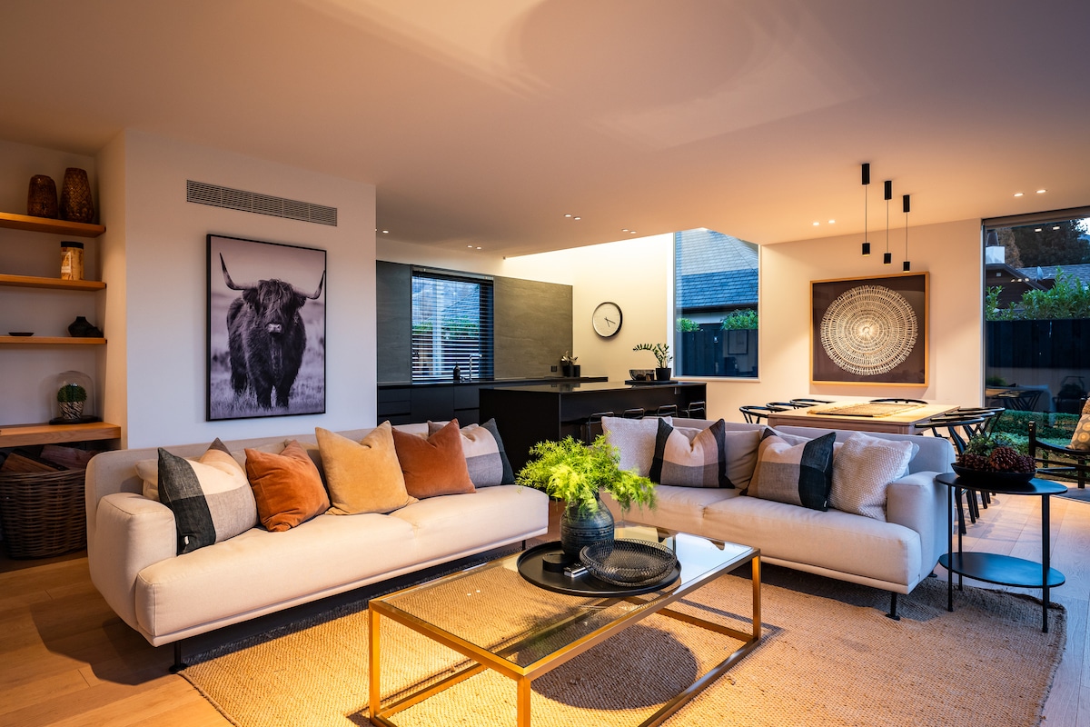 A modern living area features a light-colored sofa adorned with a variety of cushions, arranged around a glass coffee table. A large artwork of a highland cow hangs on the wall, with a blend of indoor and outdoor light illuminating the space.