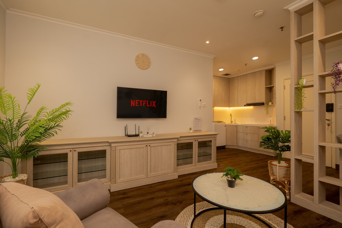 A modern living area showcases a wall-mounted smart TV displaying Netflix. Soft beige furnishings complement light wooden cabinetry, while a round table sits centrally, adorned with a small potted plant. A green indoor plant adds a touch of nature, enhancing the overall atmosphere.