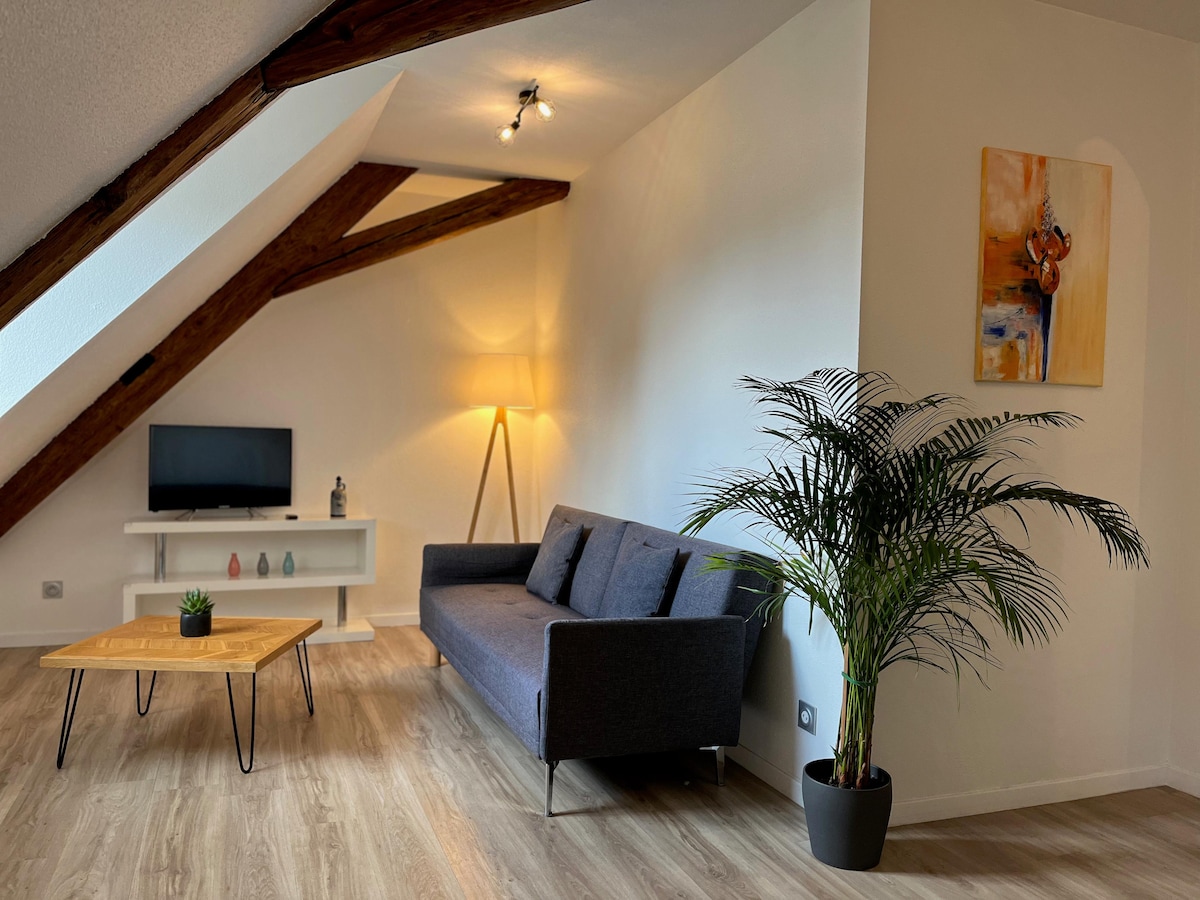 A cozy living space features exposed wooden beams and a comfortable sofa. A wooden coffee table sits in front of the sofa. A flat-screen TV is mounted on the wall above a minimalist shelf. A standing lamp provides soft lighting, and a potted plant adds a touch of greenery.