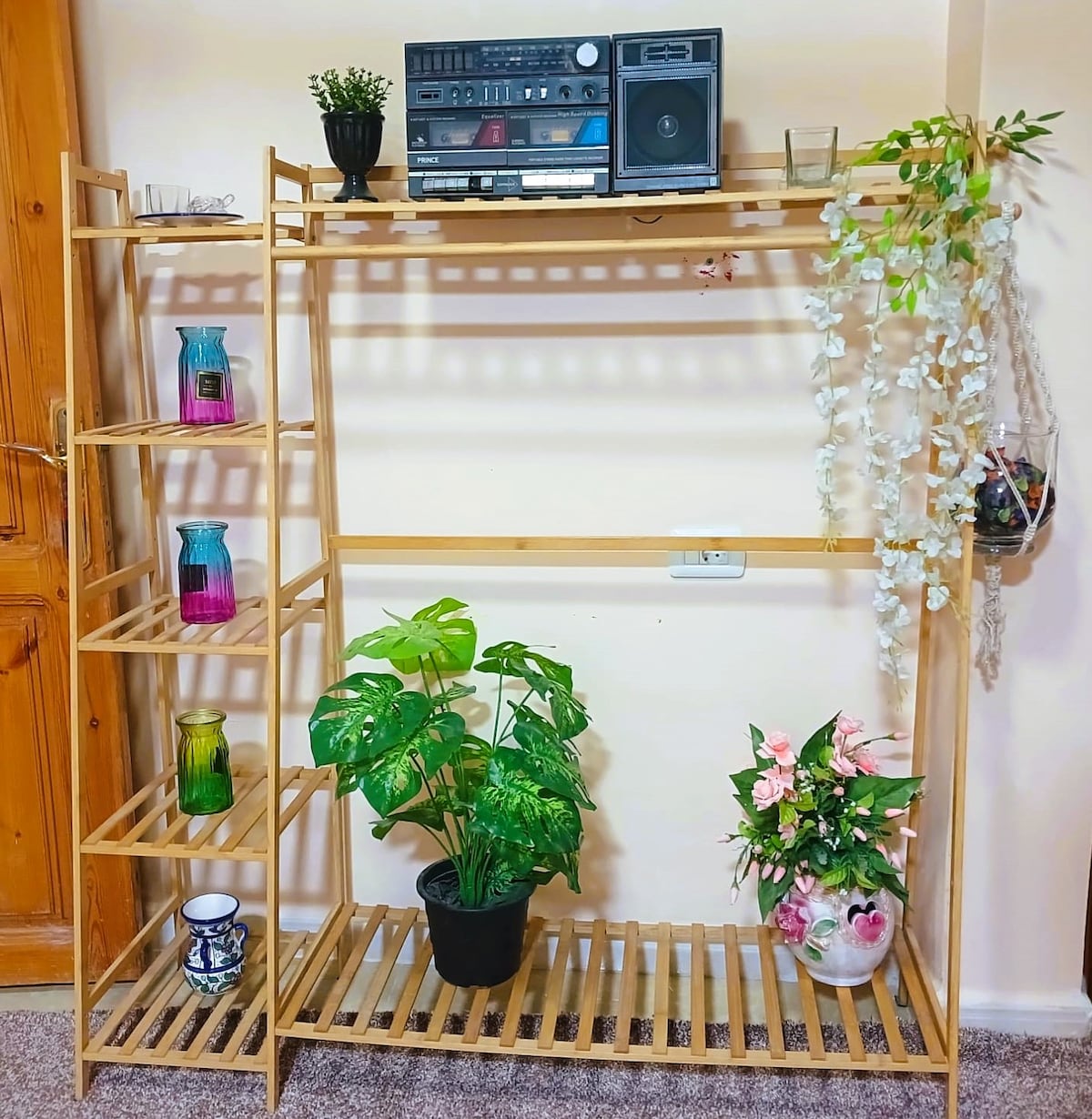 A wooden shelving unit displays various decorative items, including colorful glass vases and lush green plants. A vintage stereo rests atop the shelves, while hanging plants add a touch of natural beauty to the arrangement.