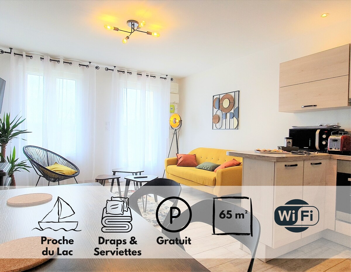 A spacious living area showcases modern furniture, including a yellow sofa and a dining table. Large windows allow natural light to illuminate the room, and a plant adds a touch of greenery. The open kitchen is equipped with appliances, providing a functional space for cooking.