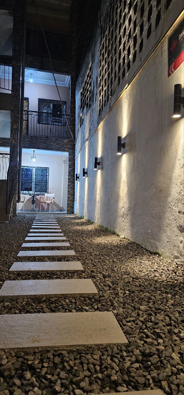 A pathway composed of rectangular stone tiles is laid over a bed of small gravel. Soft lighting fixtures are mounted on the wall, illuminating the space. In the background, a glimpse of outdoor seating is visible, framed by a rustic wall and surrounding greenery.
