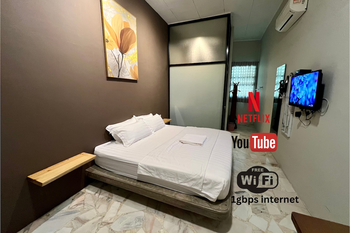 An intimate sleeping area features a spacious bed adorned with fresh linens. A wall-mounted television displays access to Netflix and YouTube. Natural light filters through a window, while a piece of wall art adds a subtle touch to the room's design.