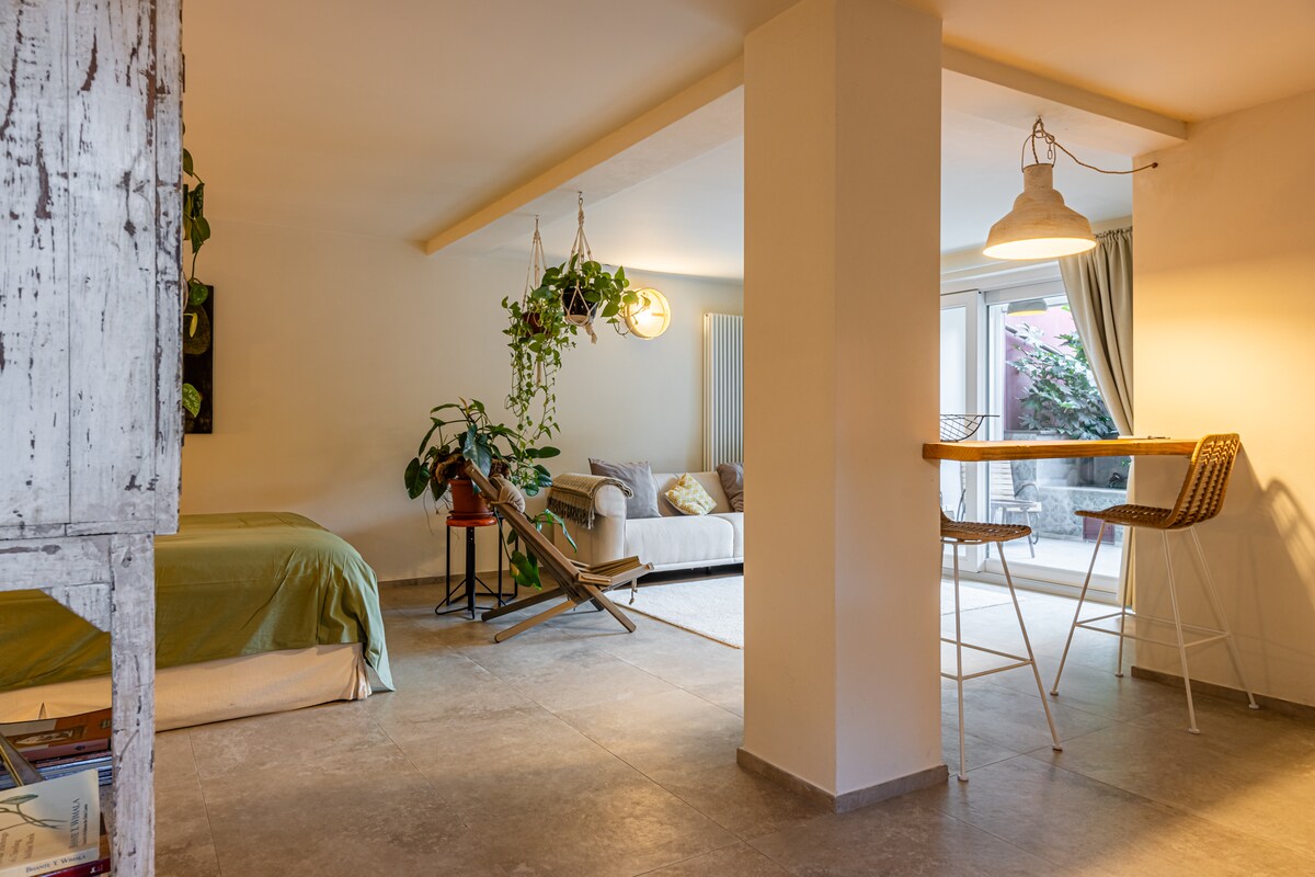 An open-plan living space is depicted, featuring a cozy sleeping area with a green blanket draped over the bed. A comfortable sofa sits beside a large window, while a small dining space with bar stools is in the foreground, complemented by hanging plants.