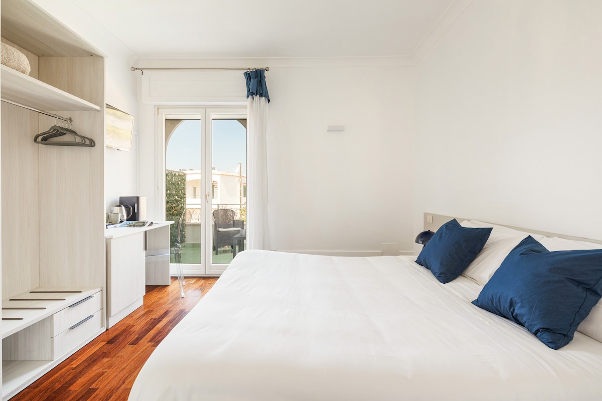 A bright and airy bedroom features a spacious bed with white linens and navy blue accent pillows. A small desk and chair are positioned beside a wardrobe, and a sliding door leads to a private balcony with outdoor seating.