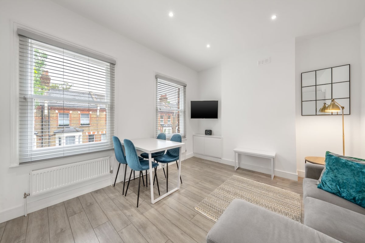 A modern open-plan living area features sleek furnishings and light wood flooring. A rectangular dining table with four blue chairs sits in natural light from large windows, while a comfortable gray sofa offers a relaxed seating option. A wall-mounted TV is positioned across from the dining area.
