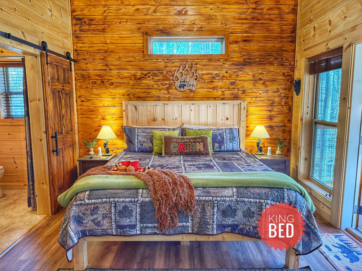 A cozy bedroom features a wooden king bed, adorned with textured bedding and decorative pillows. Soft lamps illuminate the rustic wood walls, complementing the warm tones. Large windows allow natural light to fill the space, providing a connection to the surrounding forest.