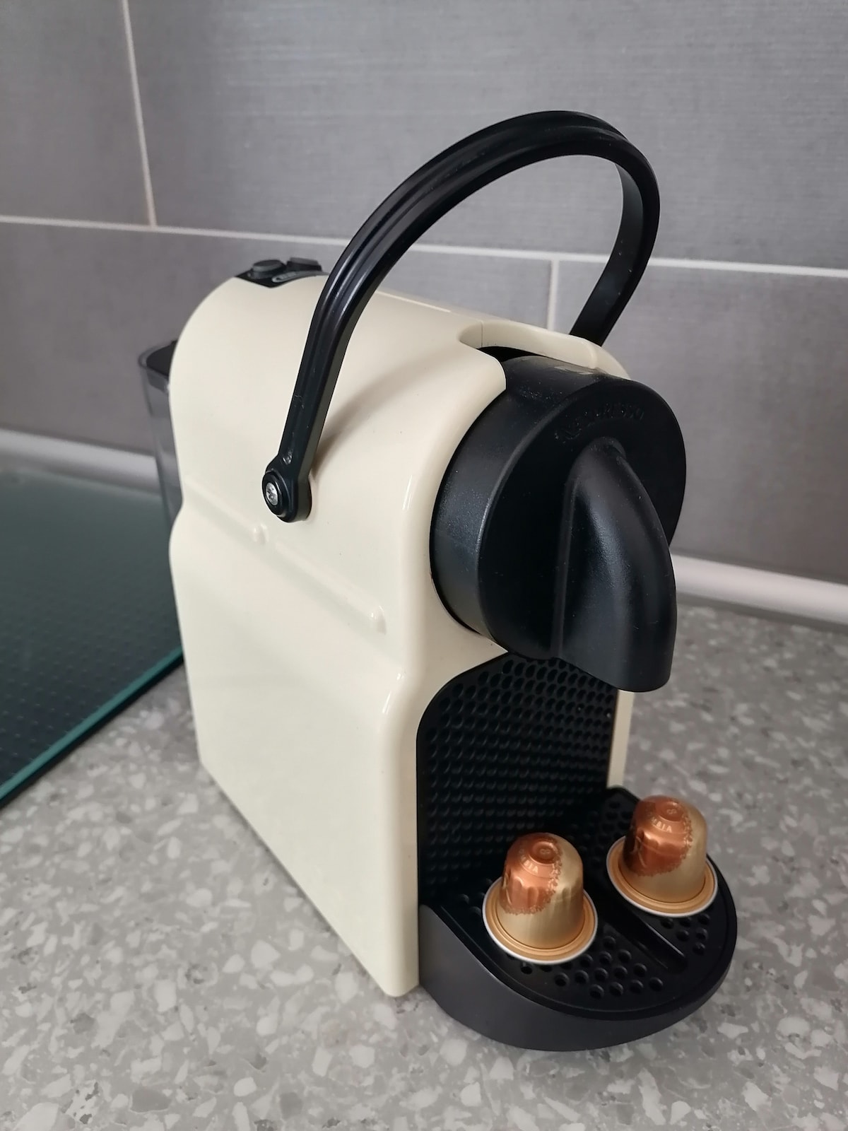 A compact coffee machine is positioned on a textured countertop. The machine features a cream-colored body with a black base and a handle. Two coffee capsules are placed in the designated slots at the front, ready for use.