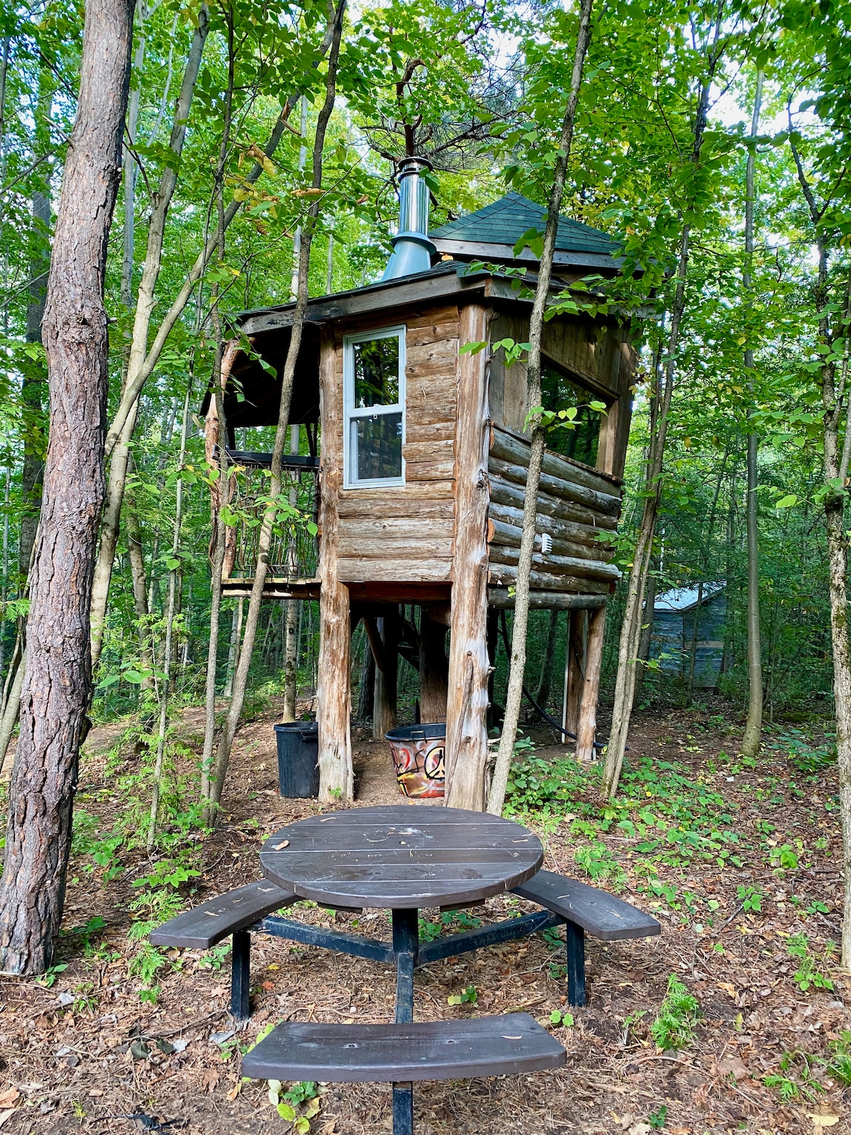 A treehouse cabin is elevated above the ground, constructed from logs, with a chimney visible on the roof. Below, a circular picnic table made of dark wood is surrounded by a natural forest setting, with greenery and tree trunks surrounding the area.