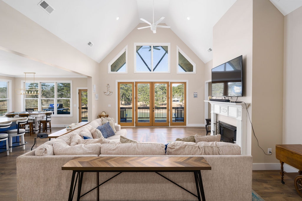 A spacious living area features a vaulted ceiling with large windows allowing natural light. The room includes a plush sectional sofa and a stylish coffee table, while a large TV is mounted above a decorative fireplace. Views of the outdoor patio are visible through the glass doors.