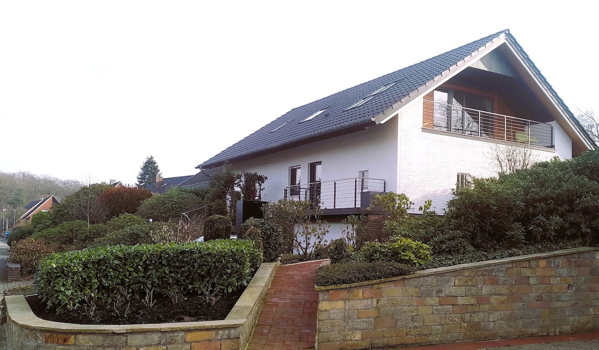 The exterior of a spacious house is shown, featuring a sloped roof and multiple windows. A pathway leads to the entrance, surrounded by well-maintained greenery and landscaped shrubs. A balcony can be seen above, providing an inviting outdoor space.