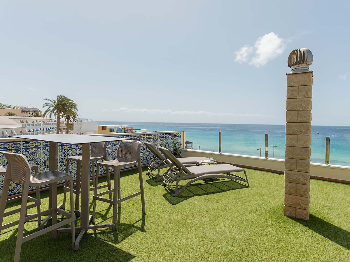 A rooftop terrace is visible with a green artificial lawn, featuring a set of tall bar stools and a table. Two sun loungers are positioned for relaxation, offering unobstructed views of the ocean and sky, with a coastal ambiance evident in the background.