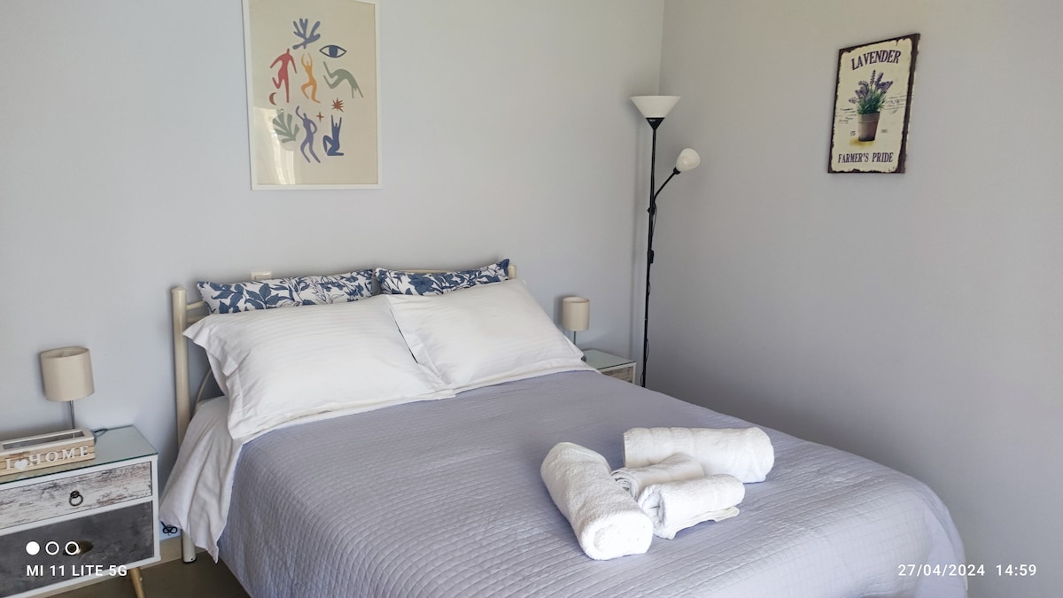 A bright sleeping area features a spacious bed with crisp white bedding and decorative pillows. Two side tables are placed beside the bed, each accompanied by a lamp. A piece of wall art adds a touch of color, while a simple floor lamp stands nearby.