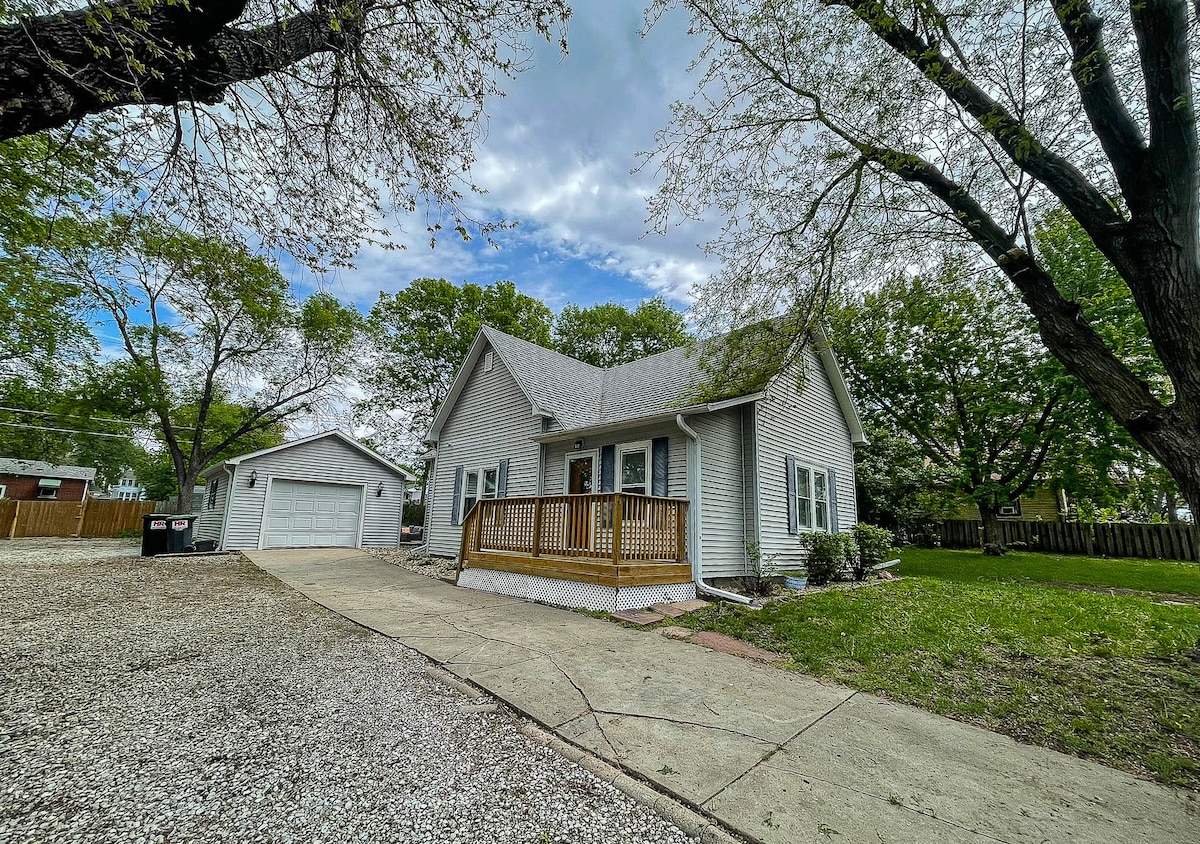 The exterior of the ranch home features a light-colored facade with a covered front porch. A gravel driveway leads to a two-car garage, bordered by green lawn and trees. The surrounding grounds present a combination of paved and grassy areas.