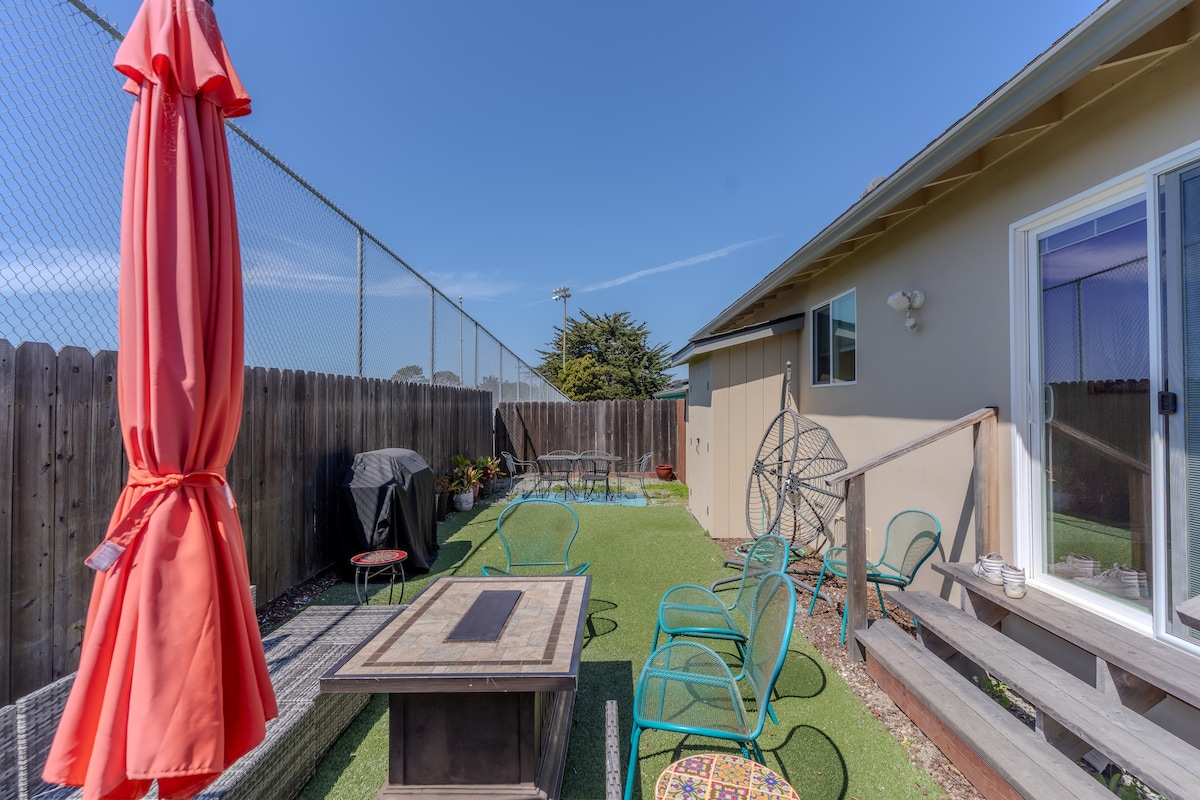 An outdoor space features a grassy area with a wooden fire pit table at its center. Surrounding seating is composed of several green chairs, and a red umbrella provides shade. A barbecue grill is placed against the wooden fence along with decorative plants.