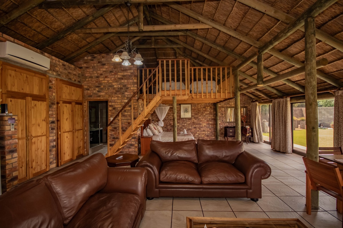 An open-plan living area showcases a high thatched ceiling supported by wooden beams. Two leather sofas are arranged invitingly, facing a central wooden table. A loft area with two single beds is visible above. Large doors lead to an outdoor space, bringing in natural light.