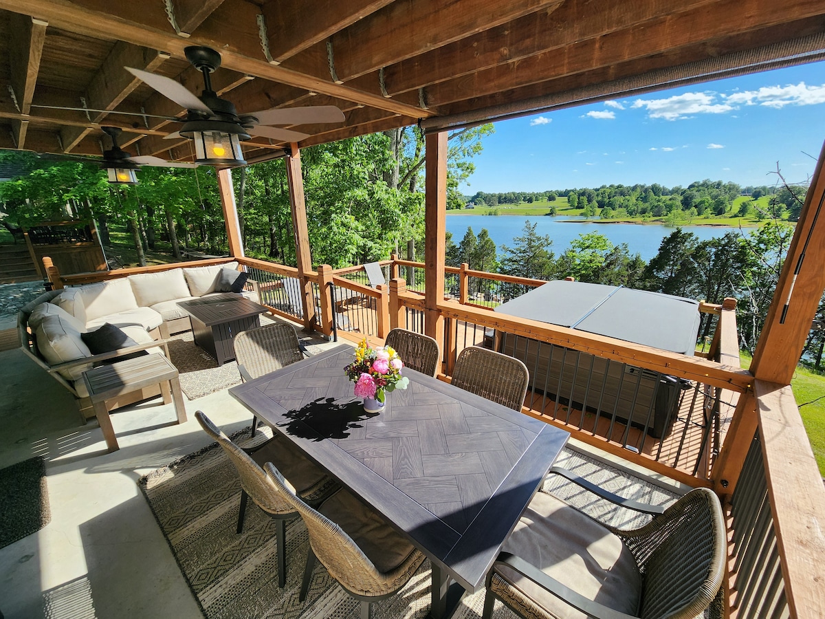 An outdoor seating area features a large table with comfortable chairs, accompanied by a sectional sofa adorned with soft cushions. A hot tub is positioned nearby, with a scenic view of the lake and trees visible in the background. Natural light enhances the inviting atmosphere.