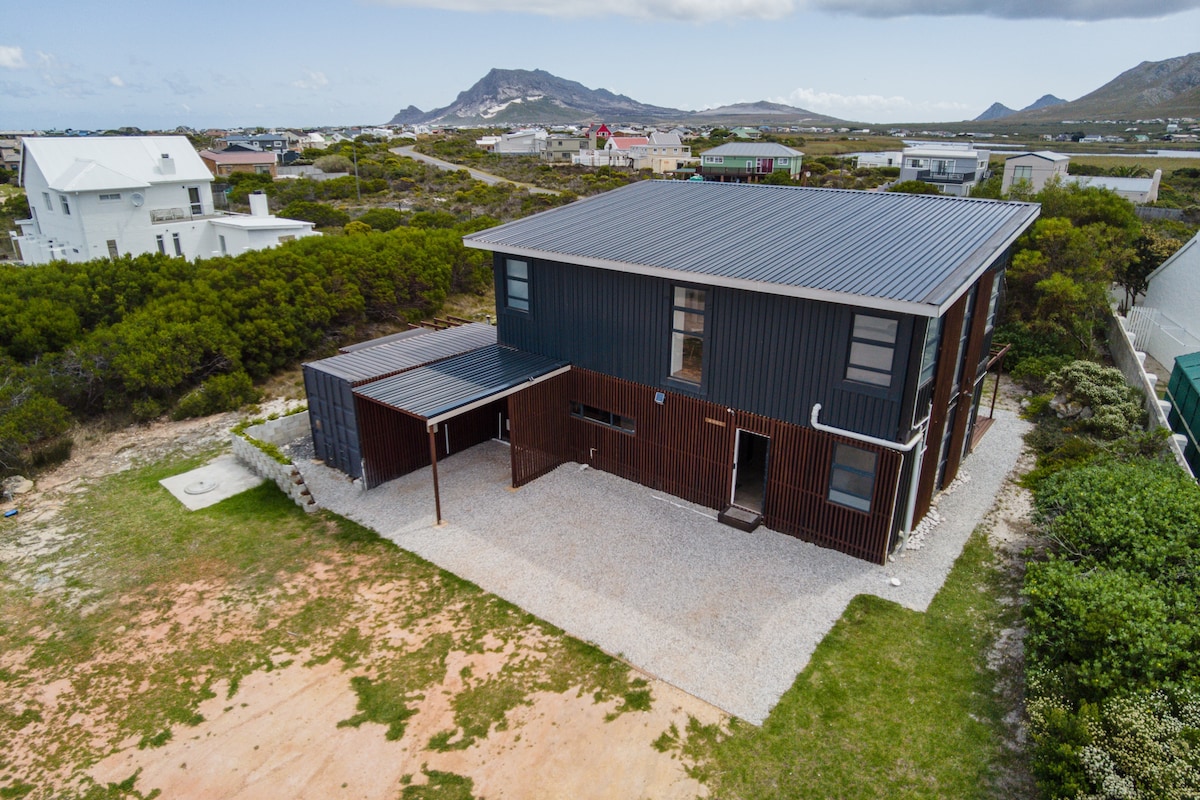 An aerial view of the eco-friendly container home showcases a spacious layout surrounded by greenery. The two-story house features a modern exterior with a dark facade and a metal roof. Nearby, a gravel area provides off-street parking, with a scenic mountain backdrop.