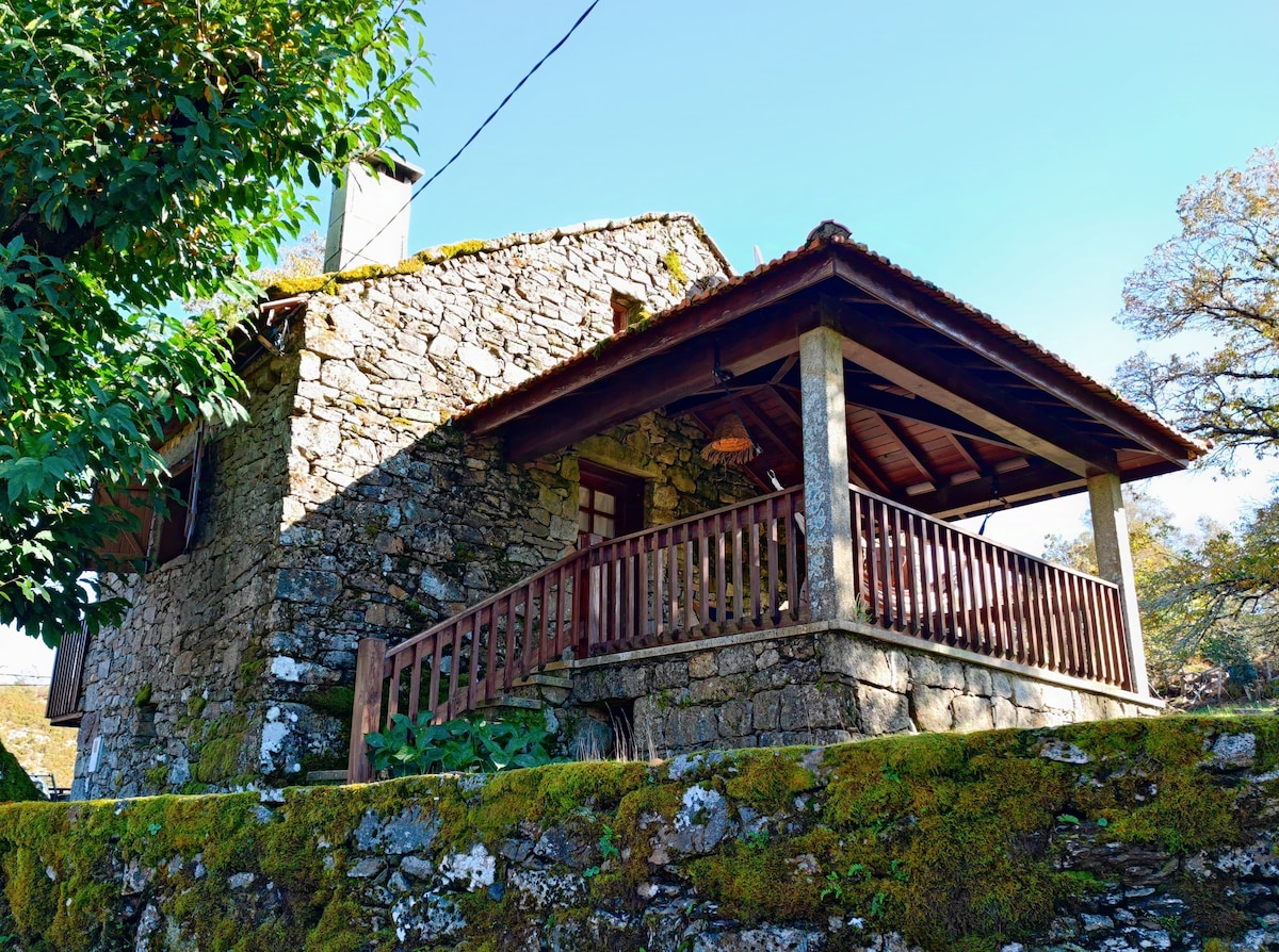 An inviting stone cottage with a wooden deck is situated under clear blue skies. The structure features rustic charm, complemented by a sloping roof and overhanging eaves. Surrounding greenery adds to the serene atmosphere, while moss-covered stones form a natural boundary.