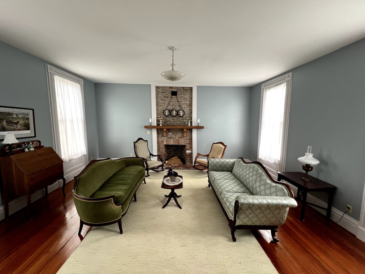 The living room features two vintage sofas, including a green one and a light blue patterned one, arranged around a small coffee table. A brick fireplace with decorative accents is centered on the wall, and large windows provide natural light, enhancing the room's inviting atmosphere.