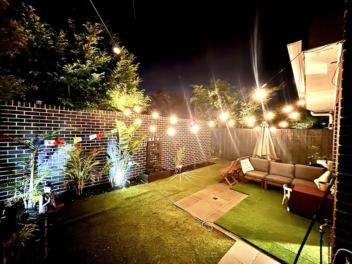 A cozy outdoor space is illuminated by string lights, creating a warm ambiance in the evening. Lush green plants are scattered throughout the area, with a comfortable seating arrangement made of a spacious sofa and side tables nestled on an artificial lawn.