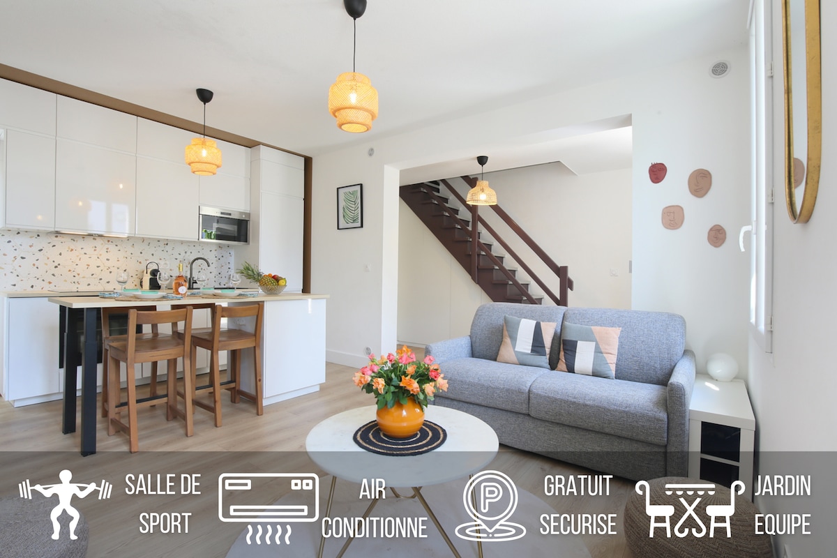 An open living area features a modern kitchen with light cabinetry and pendant lighting above a dining space. A comfortable gray sofa is positioned beside a flowering centerpiece on a circular table, while a staircase leads to an upper level in the background.