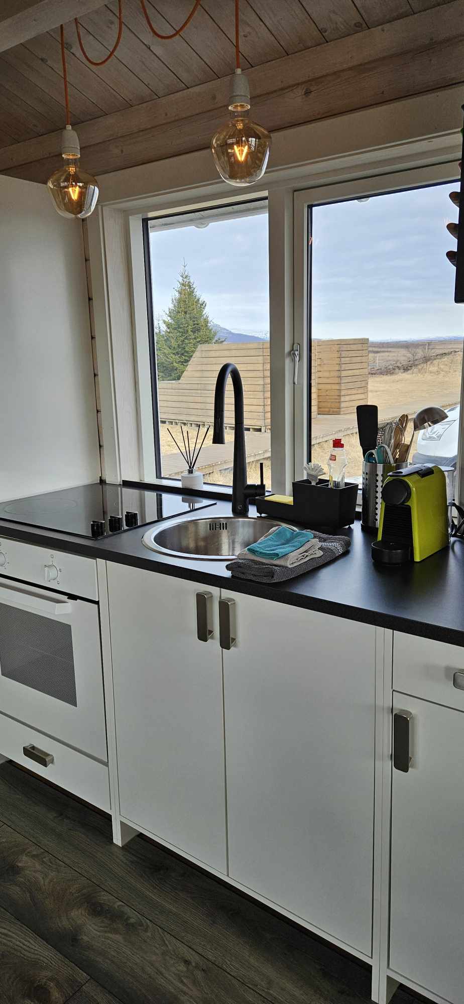 A compact kitchen area is illuminated by natural light from a large window. Modern appliances include a fridge and oven, with a sleek black countertop housing a sink and various kitchen utensils. Subtle decorations complement the functional layout, creating an inviting cooking space.