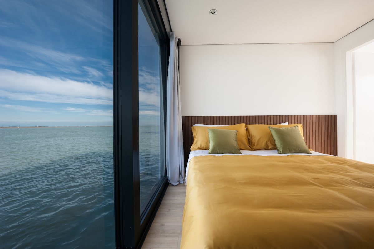 A bright bedroom features a spacious bed dressed in golden linens and accented with green pillows. Large glass windows allow stunning water views, creating a seamless connection to the outside. Soft natural light enhances the serene atmosphere of the room.