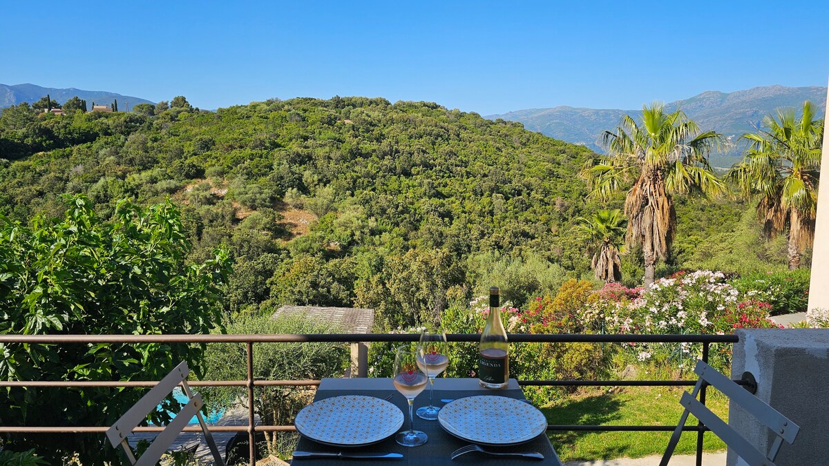 A scenic terrace is viewed, featuring a dining table set for two with plates and a bottle of wine. Lush green hills and palm trees are visible in the background, creating a peaceful outdoor space surrounded by nature.