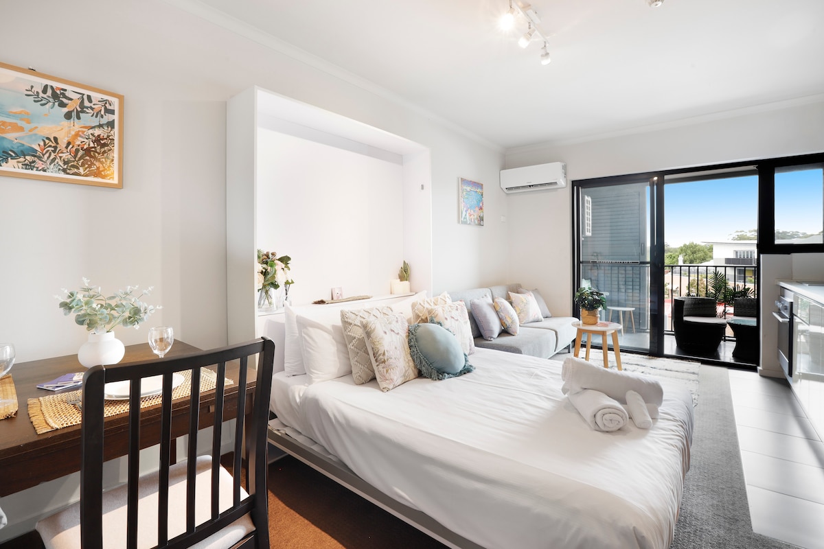 A bright studio apartment showcases a fold-out Murphy bed with crisp linens, adjacent to a cozy lounge area featuring a sectional sofa. A dining table with two chairs is placed nearby, while large sliding doors lead to a balcony with city views.