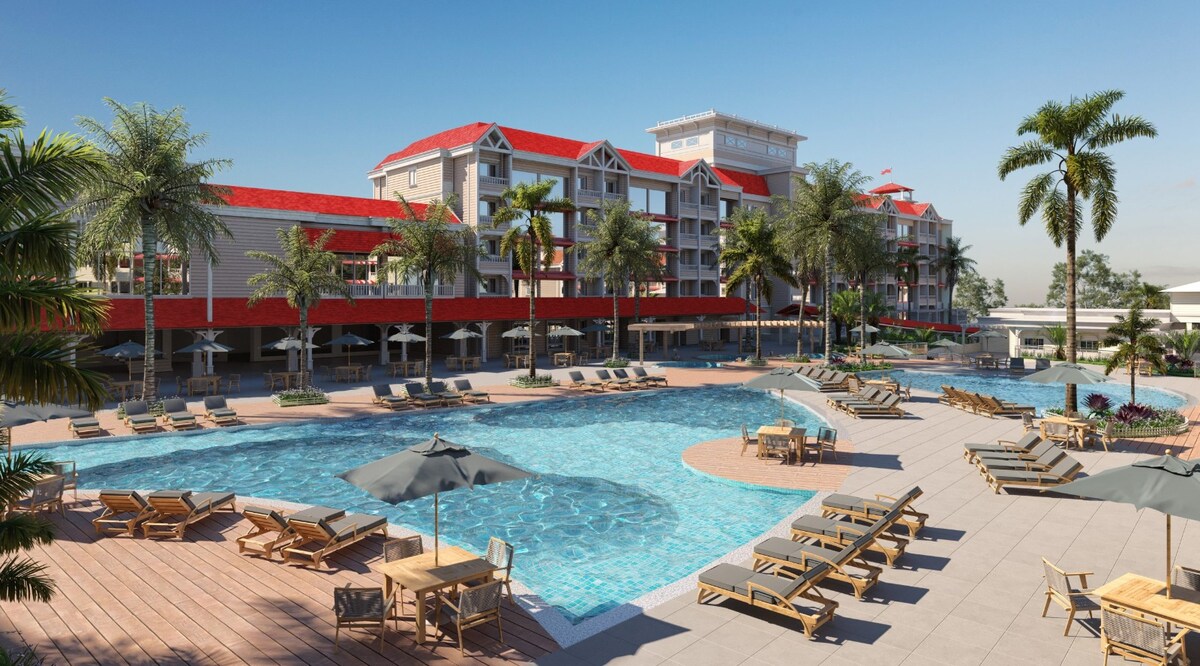 The resort complex features a large outdoor pool surrounded by sun loungers and umbrellas. Palm trees provide shade, and the building's red-roofed architecture adds charm to the setting. The space invites relaxation and enjoyment for guests of all ages.