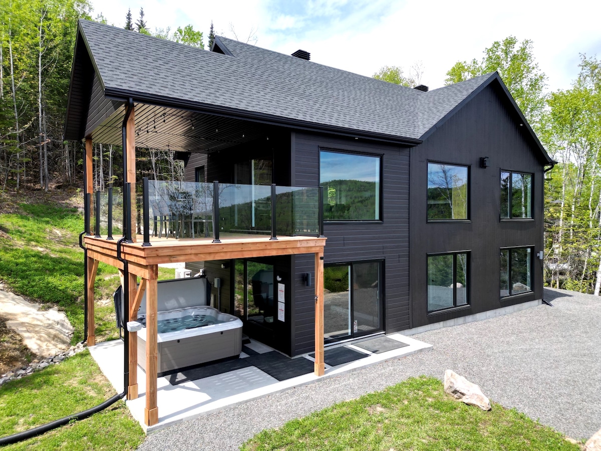 The exterior of a modern chalet features a dark wooden facade with large windows. A spacious deck is visible above the covered hot tub area. Green grass surrounds the property, and gravel paths lead to the entrance, providing a serene and natural setting.