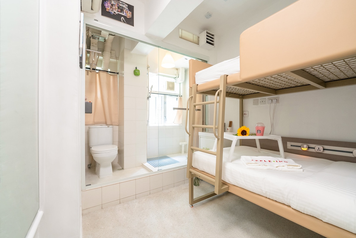 A modern room featuring a bunk bed and a separate bathroom area visible through a glass divider. The bathroom includes a shower, toilet, and basic amenities. Natural light enters through a window, highlighting the simple decor and minimalistic style.