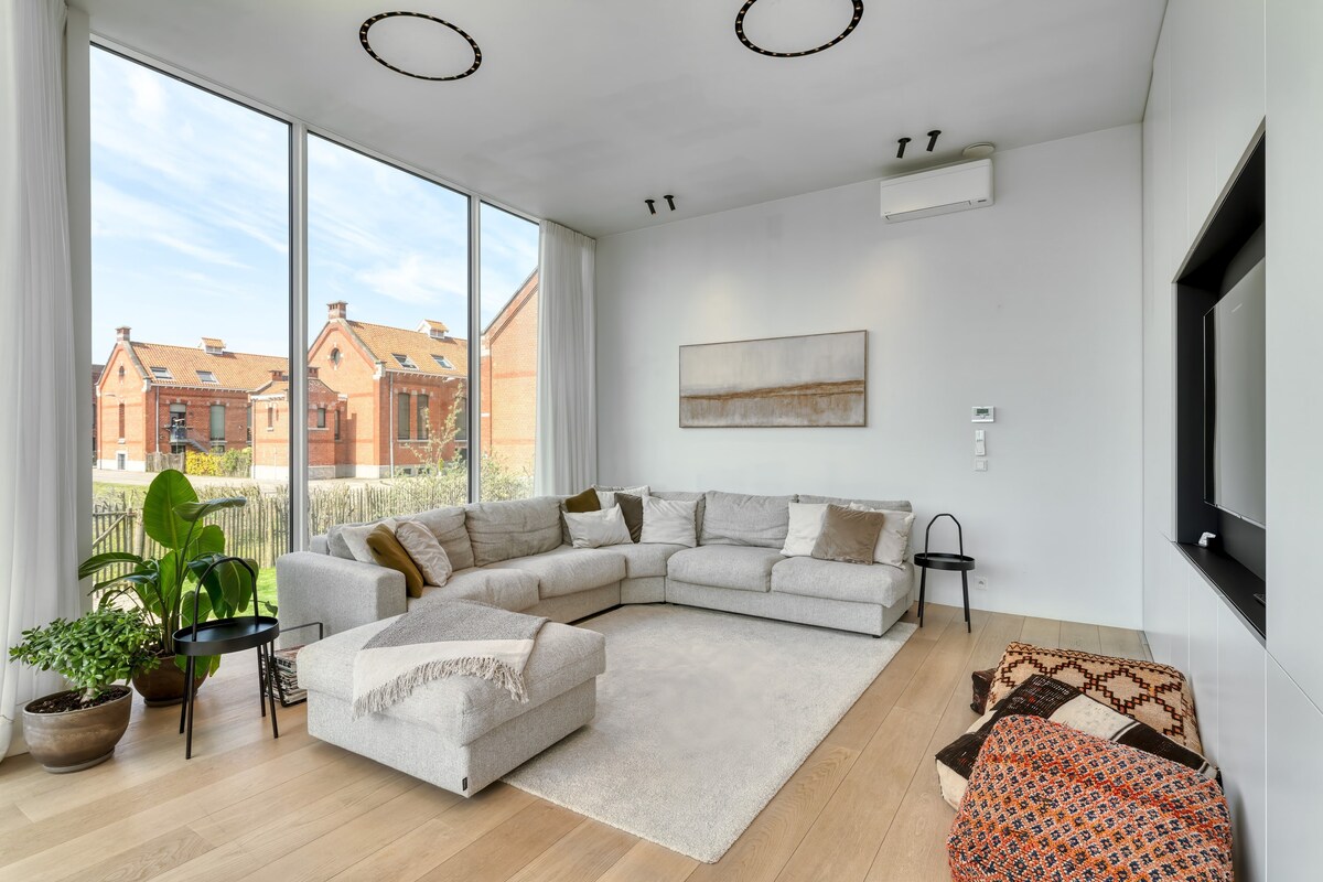 A spacious living area is featured, showcasing a large sectional sofa positioned around a soft area rug. Tall windows allow natural light to fill the room, offering views of the outdoor landscape. Decorative plants and cushions add a touch of homeliness to the contemporary space.