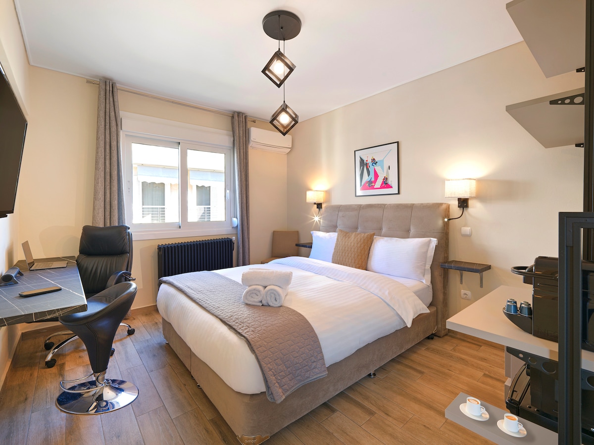 A well-appointed bedroom features a spacious bed dressed in neutral linens, accompanied by decorative pillows. A work desk and a comfortable chair are positioned near a window, allowing natural light. Soft lighting from wall sconces creates a cozy atmosphere. A contemporary art piece decorates the wall.