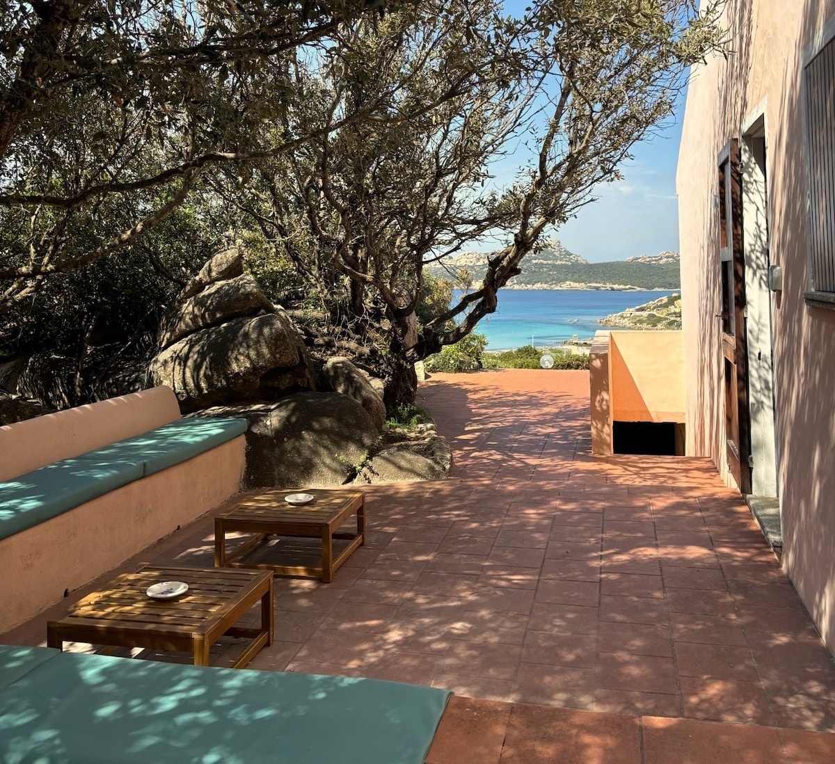 An outdoor seating area is depicted with a shaded bench and wooden tables positioned on a terracotta-tiled patio. The scene features surrounding natural stone formations and greenery, leading towards a view of the blue sea and distant islands in the background.
