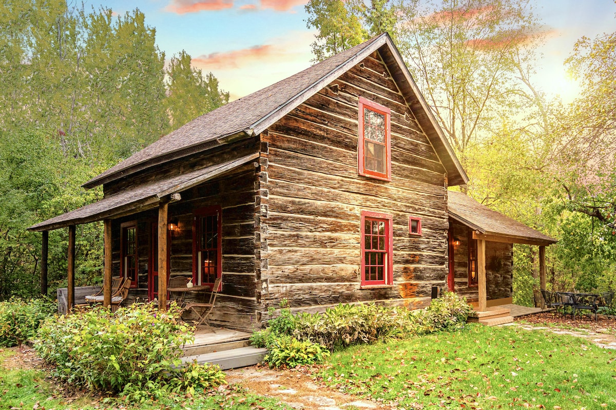 A charming log cabin features rustic wooden architecture, complemented by red window frames. Surrounded by lush greenery, the structure offers a welcoming front porch. Natural light bathes the exterior, enhancing the serene setting among the trees, with a pathway leading to the entrance.