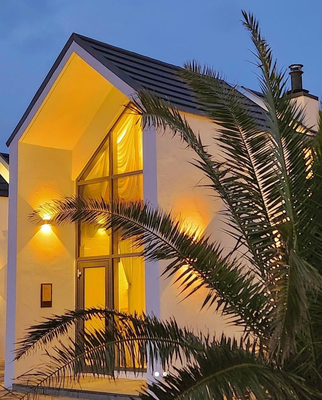 A modern structure is depicted with a peaked roof and large glass windows, allowing soft light to emanate from within. Lush palm fronds frame the foreground, enhancing the inviting ambiance created by the warm interior lighting.