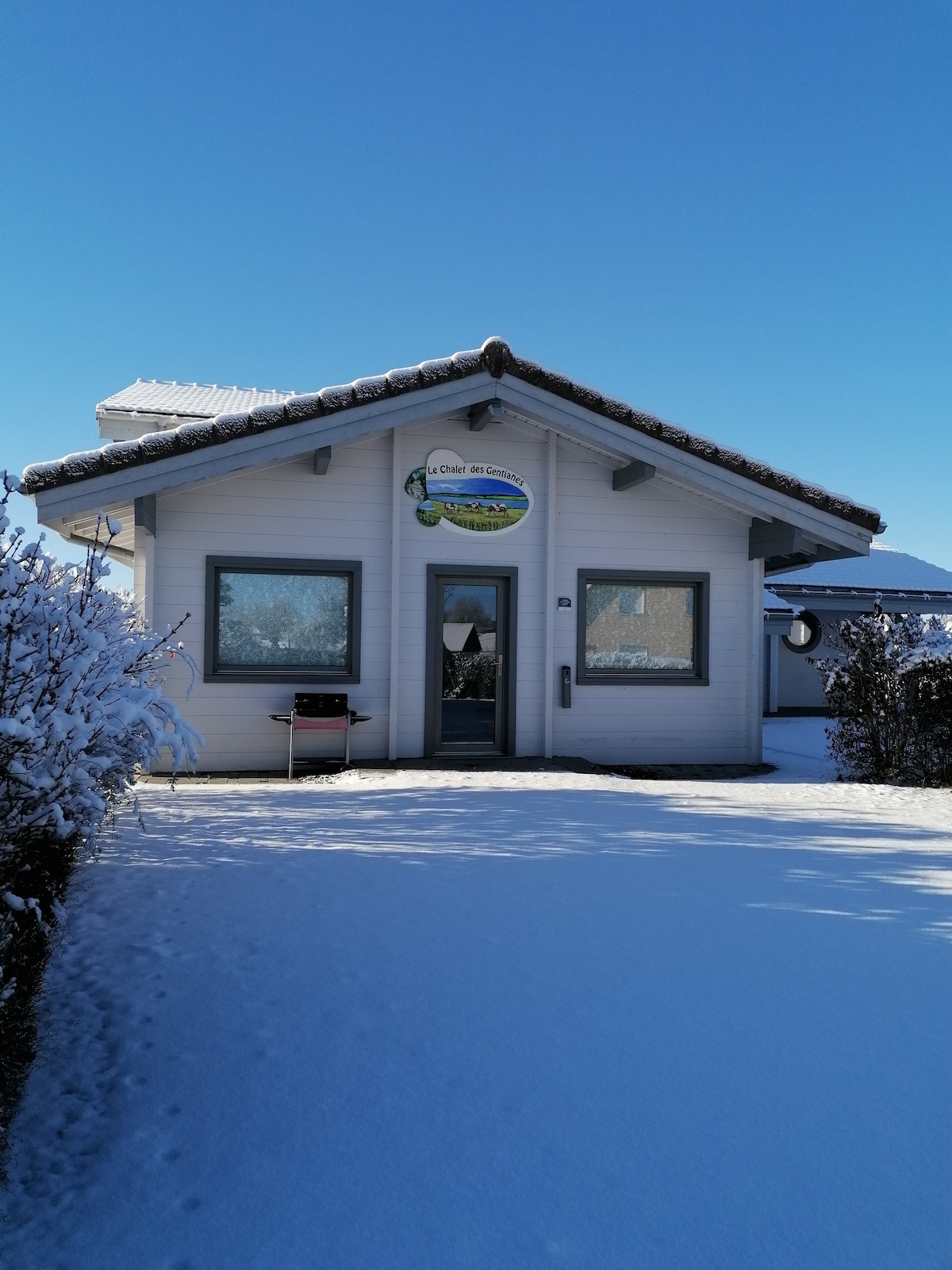 A modern chalet-style building sits beneath a clear blue sky, surrounded by a blanket of fresh snow. The entrance features large windows, a glass door, and a sign displaying the property's name. Snow-covered bushes frame the pathway leading to the entrance.